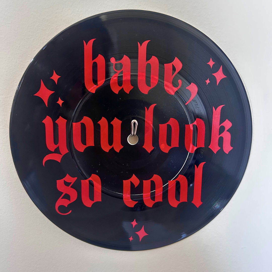 A circular black vinyl record wall decorative sign with red gothic text reading 'babe, you look so cool' and small red sparkle accents, mounted against a plain light wall.