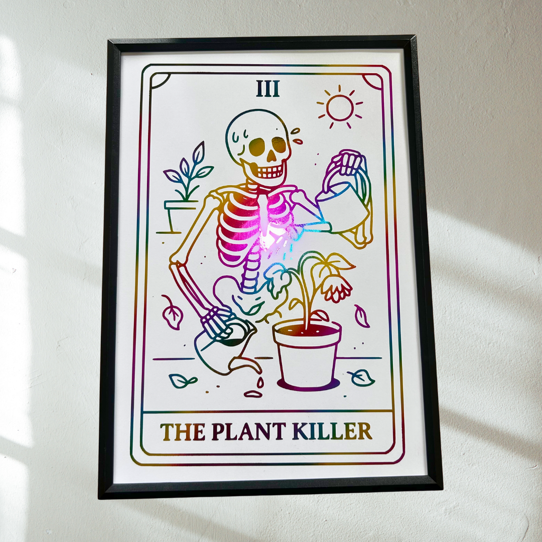 A framed art print featuring a tarot-style illustration of a skeleton gardening, labeled 'THE PLANT KILLER' with Roman numeral 'III' at the top, hanging on a white wall with soft sunlight shadows.