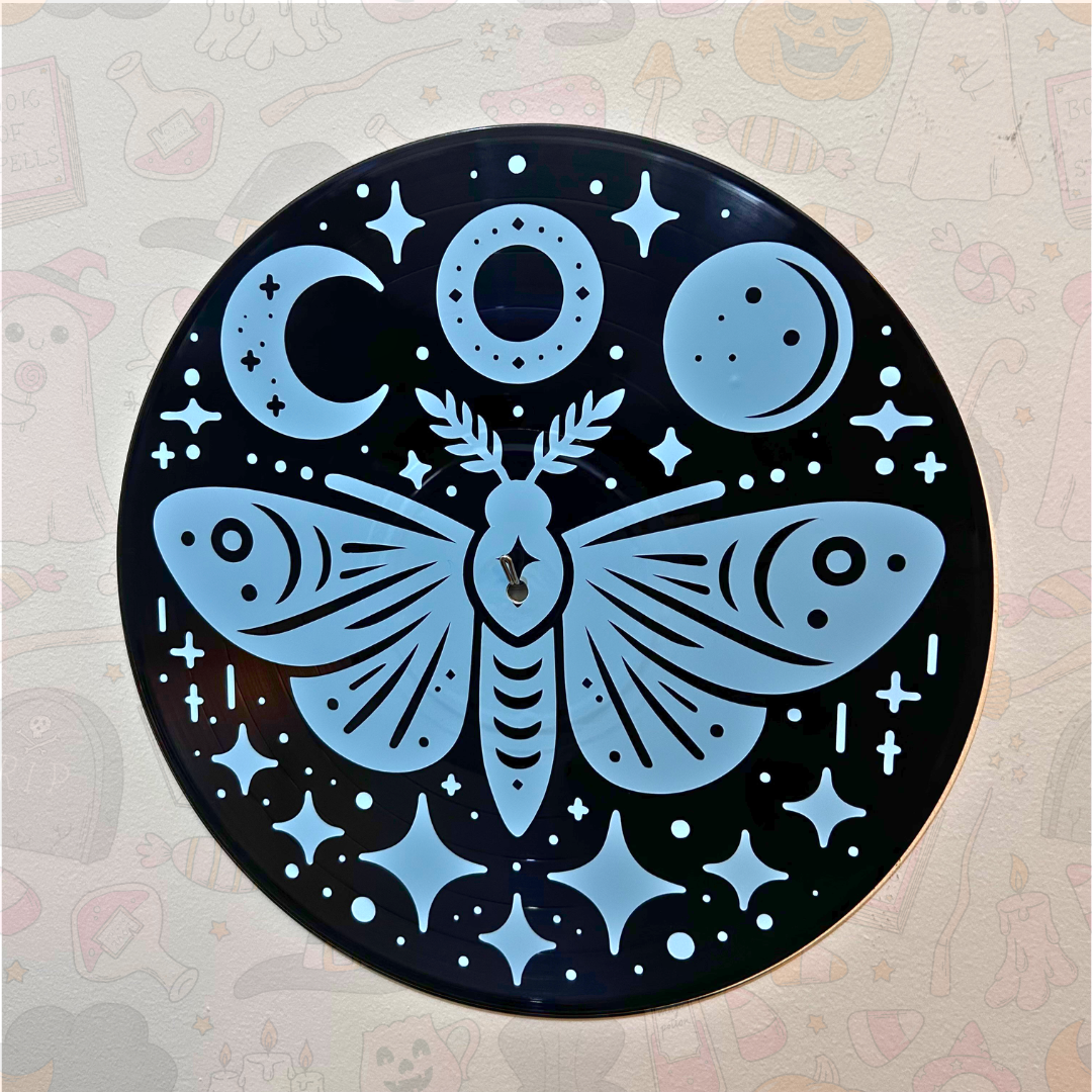 A round black wall art piece featuring a light blue celestial design with a large moth, crescent moons, stars, and dots on a patterned beige wall.