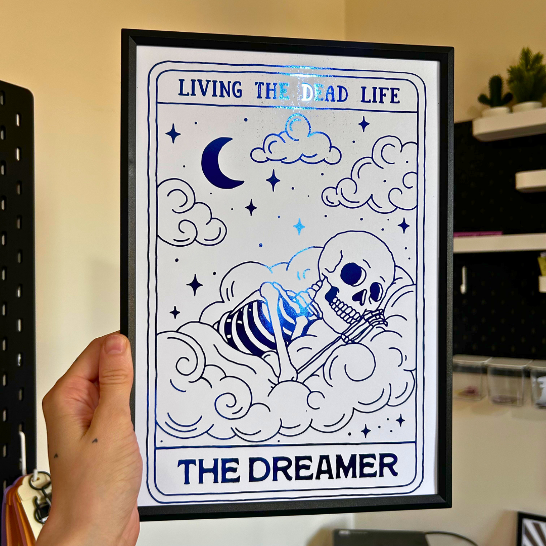 A framed art print featuring a blue and white line art illustration of a skeleton lying on clouds under a crescent moon, held by a hand. The text reads 'LIVING THE DEAD LIFE' at the top and 'THE DREAMER' at the bottom.