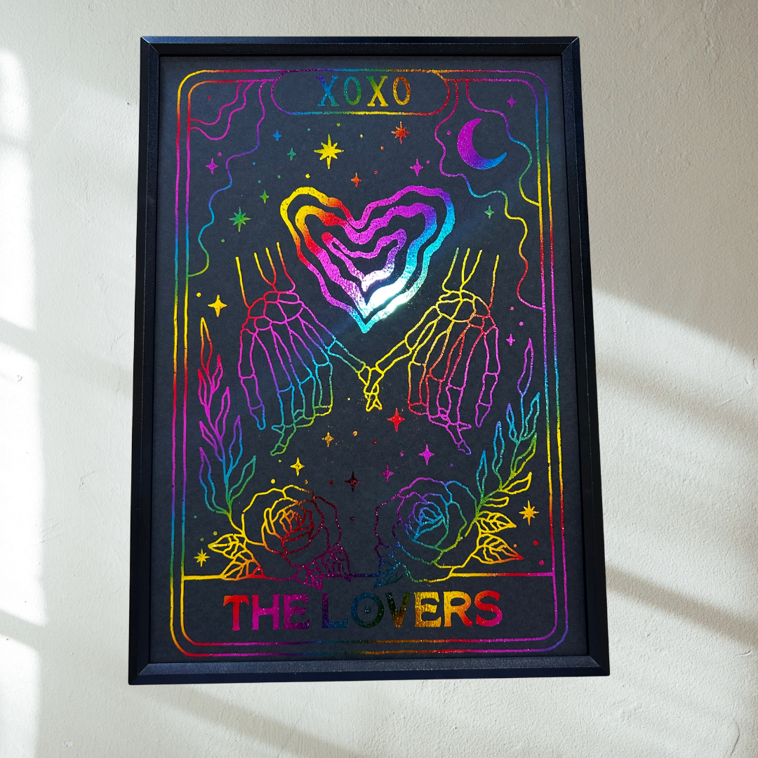 A framed tarot card art print titled 'THE LOVERS' with 'XOXO' at the top, featuring two hands holding a rainbow-colored heart on a black background, mounted on a white wall with soft sunlight shadows.