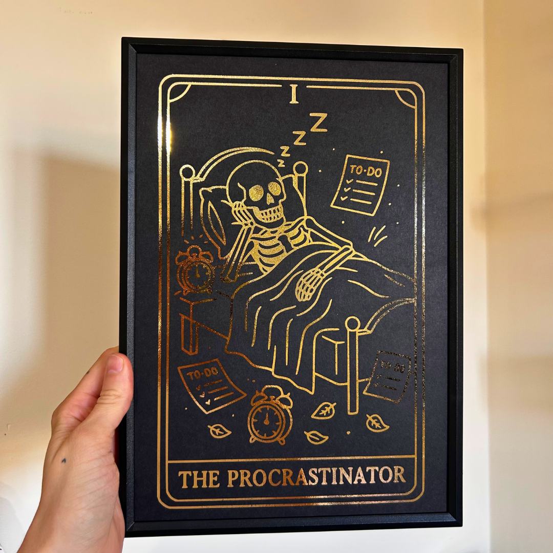 A hand holds a black-framed art print featuring a gold line-art illustration of a skeleton sleeping in bed with the text 'I Z Z' and 'THE PROCRASTINATOR' at the bottom, against a beige wall.