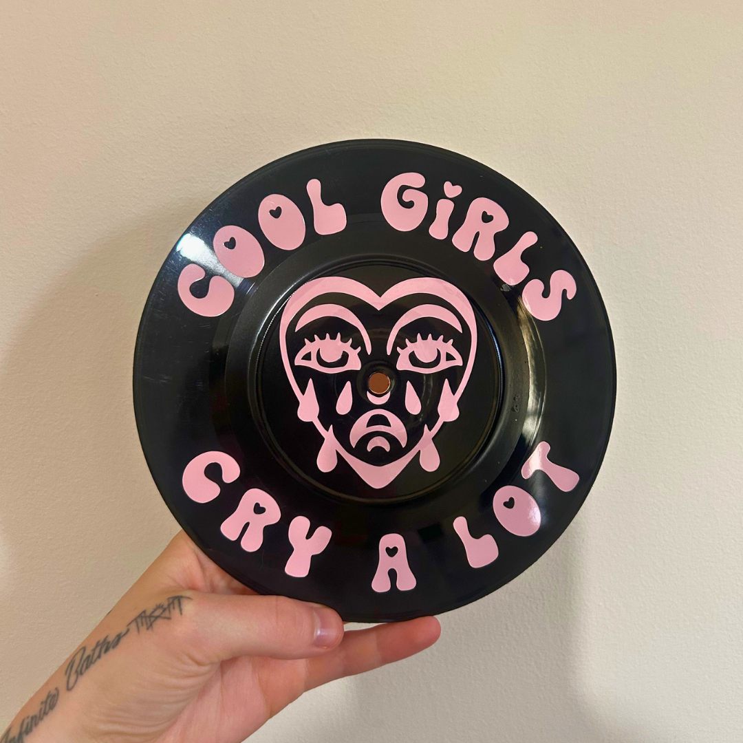 A hand holds a black circular sign made with upcycled vinyl record, with pink text reading 'COOL GIRLS CRY A LOT' and a pink stylized face in the center, against a plain beige wall.