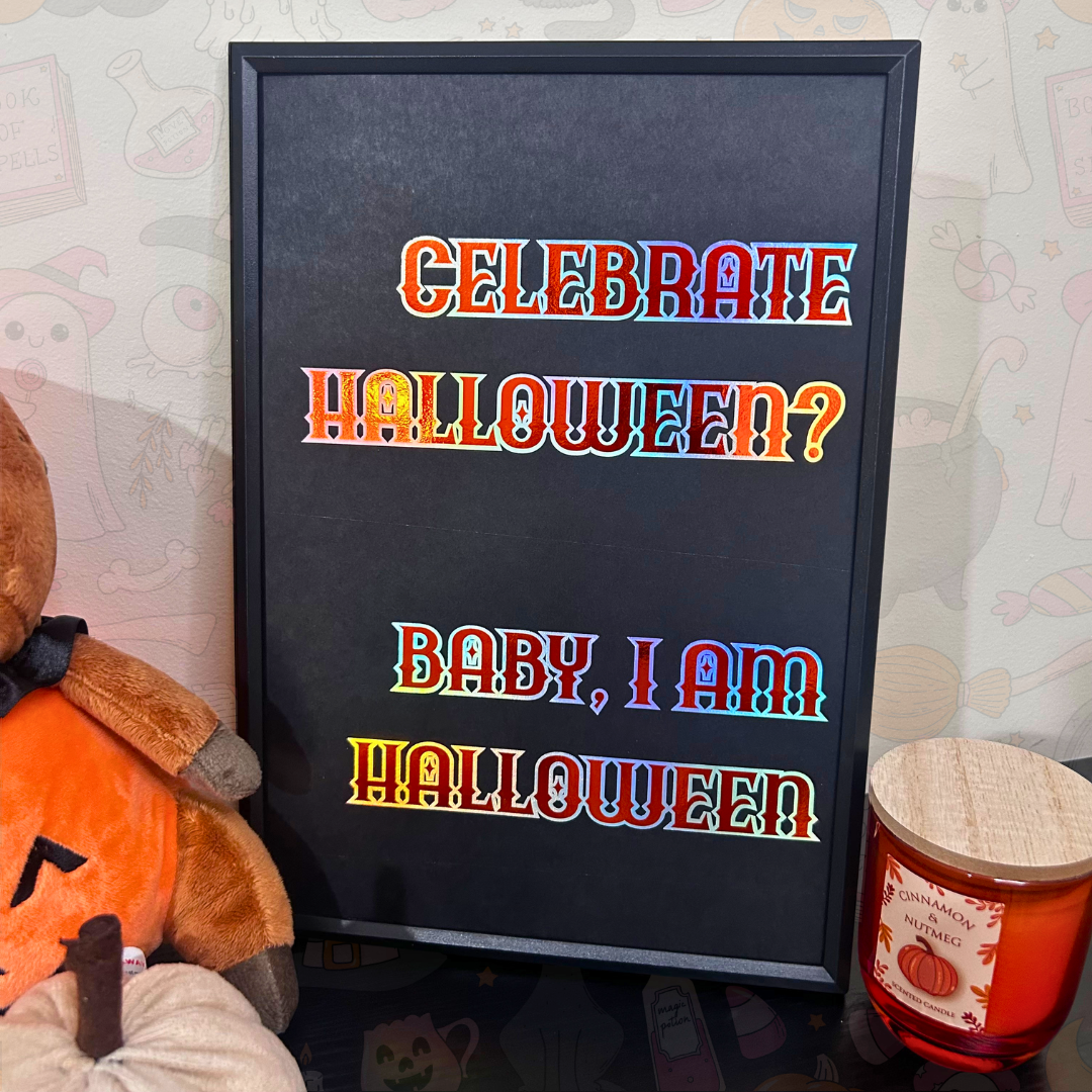 A framed Halloween-themed sign with the text 'CELEBRATE HALLOWEEN? BABY, I AM HALLOWEEN' in colorful gradient letters on a black background. The sign is styled on a tabletop next to a plush pumpkin and a candle, with a patterned wallpaper in the background.