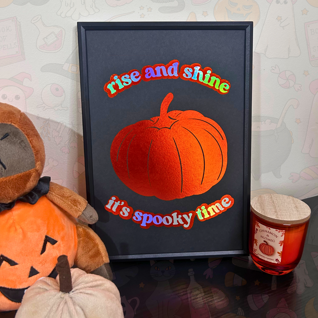 A framed Halloween art print featuring a textured orange pumpkin and the words 'rise and shine it's spooky time' in colorful letters, styled on a table next to a plush jack-o'-lantern and a candle.