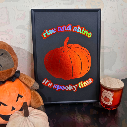 A framed Halloween art print featuring a textured orange pumpkin and the words 'rise and shine it's spooky time' in colorful letters, styled on a table next to a plush jack-o'-lantern and a candle.
