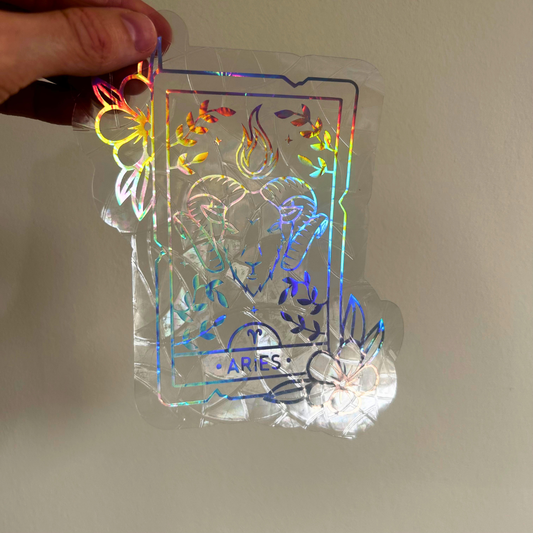 A hand holds a clear acrylic sticker featuring a holographic Aries zodiac card design with a ram illustration and rainbow iridescent foil effects, shown against a plain beige wall.