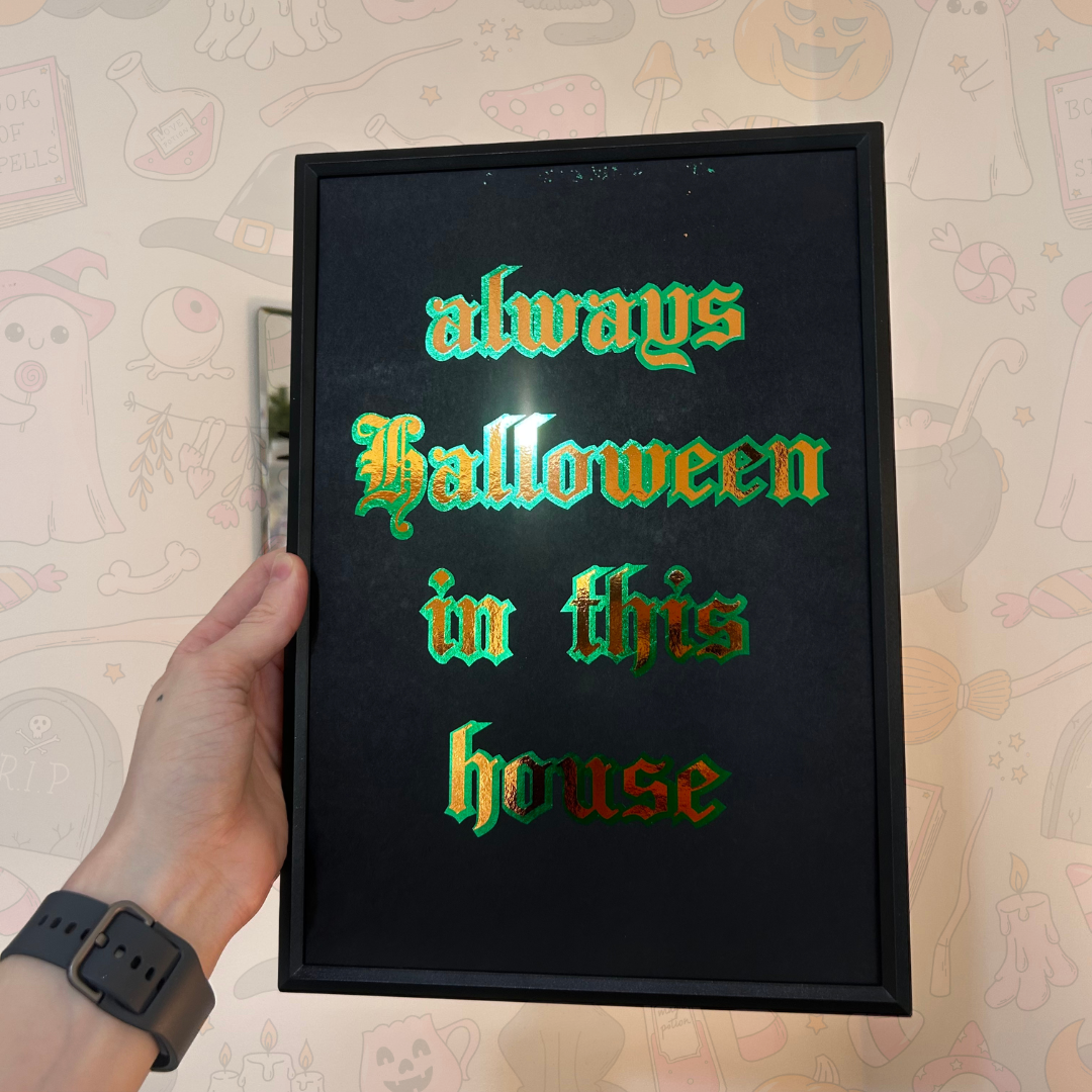 A hand holds a black rectangular framed sign with the phrase 'always Halloween in this house' in raised gothic-style lettering that shifts between green and red. The background is a light beige patterned wallpaper with faint cartoon illustrations.