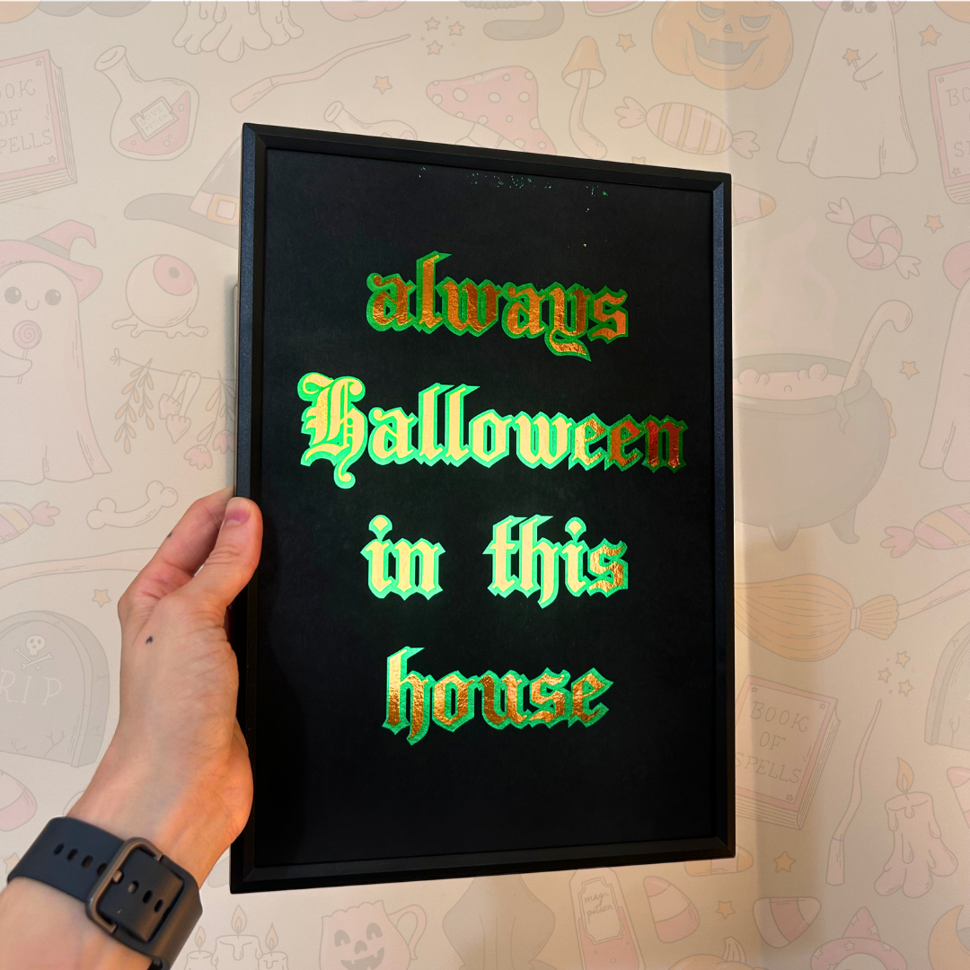 A hand holds a black-framed decorative sign with the phrase 'always Halloween in this house' in glowing green gothic lettering against a light patterned wall.