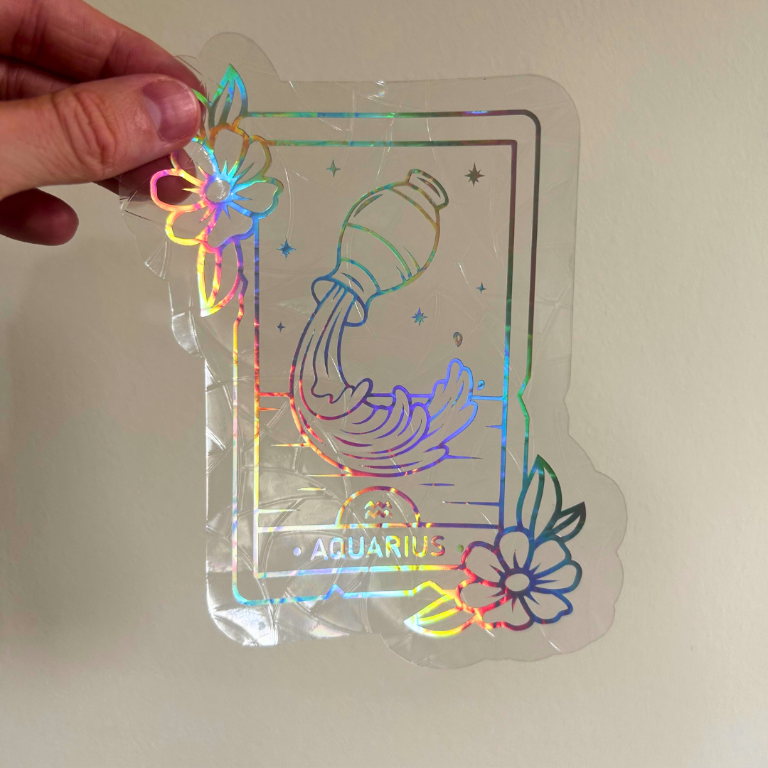 A hand holds a clear acrylic sticker featuring an Aquarius zodiac design with a holographic rainbow foil border and floral accents, set against a plain light background.