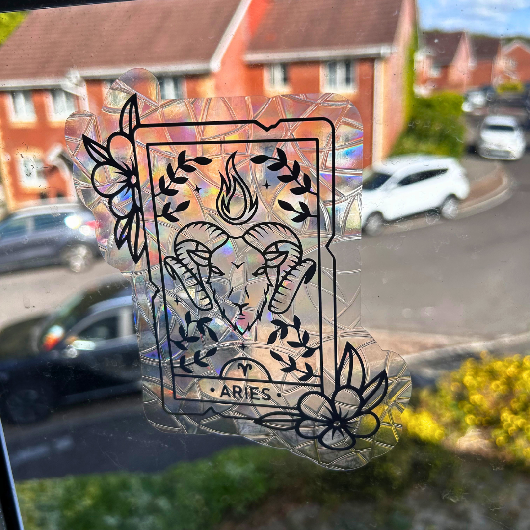 A holographic, crinkled plastic sticker with a black line art ram illustration and the word 'ARIES' at the bottom, shown attached to a window with a blurred suburban background.