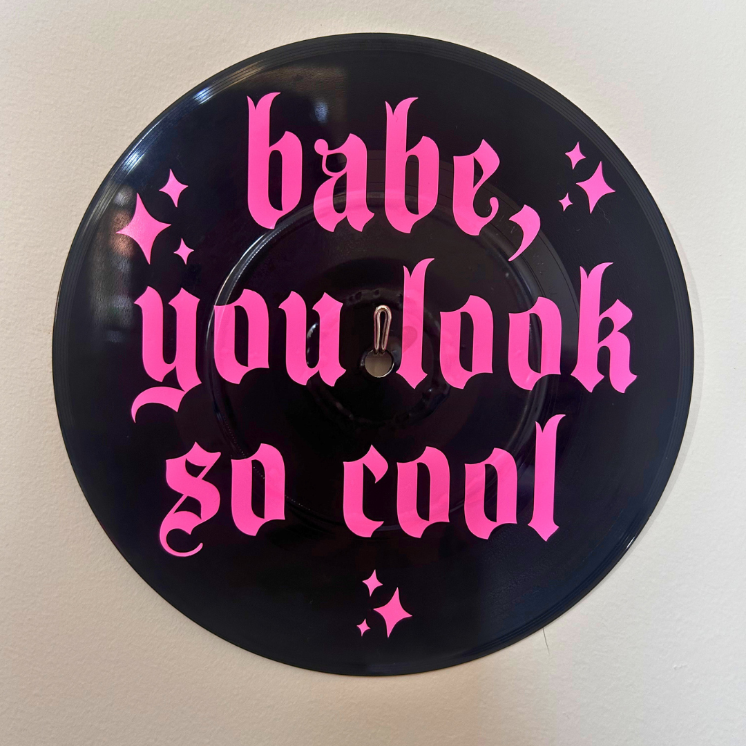 A circular black vinyl record with bright pink gothic text reading 'babe, you look so cool' and small pink sparkle accents, mounted against a plain light wall.