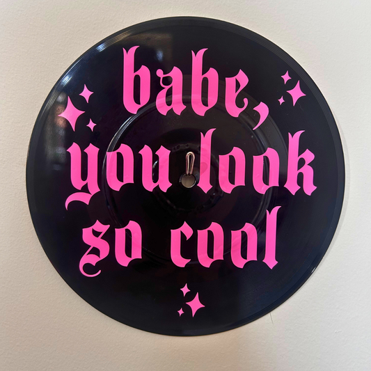 A circular black vinyl record with bright pink gothic text reading 'babe, you look so cool' and small pink sparkle accents, mounted against a plain light wall.