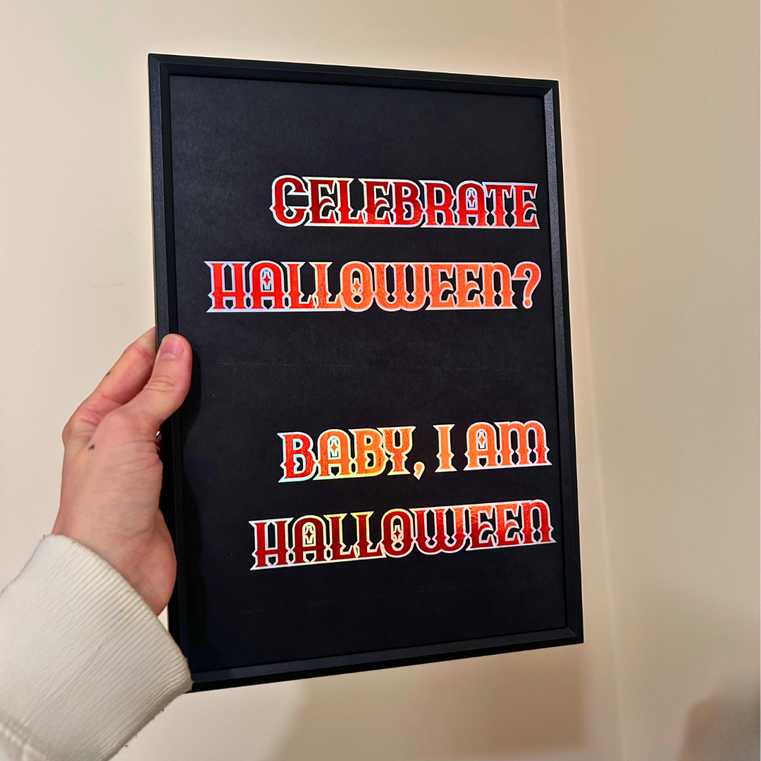 A hand holding a black framed sign with the text 'CELEBRATE HALLOWEEN? BABY, I AM HALLOWEEN' in red and orange letters with silver outlines, against a beige wall.