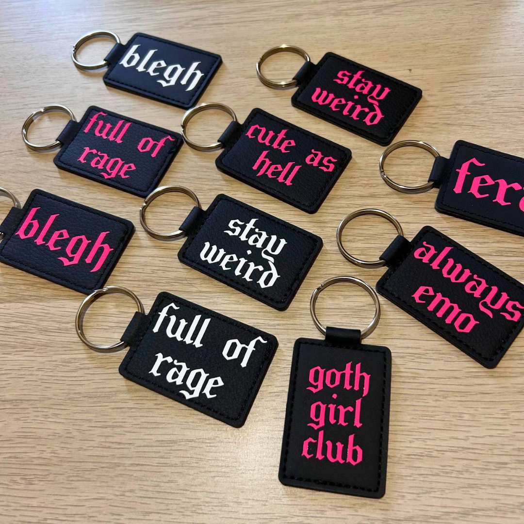 A set of black rectangular embroidered keychains with metal key rings, arranged on a light wood surface. Each keychain has a different phrase embroidered in white or hot pink, including 'blegh', 'stay weird', 'full of rage', 'cute as hell', 'feral', 'always emo', and 'goth girl club'.