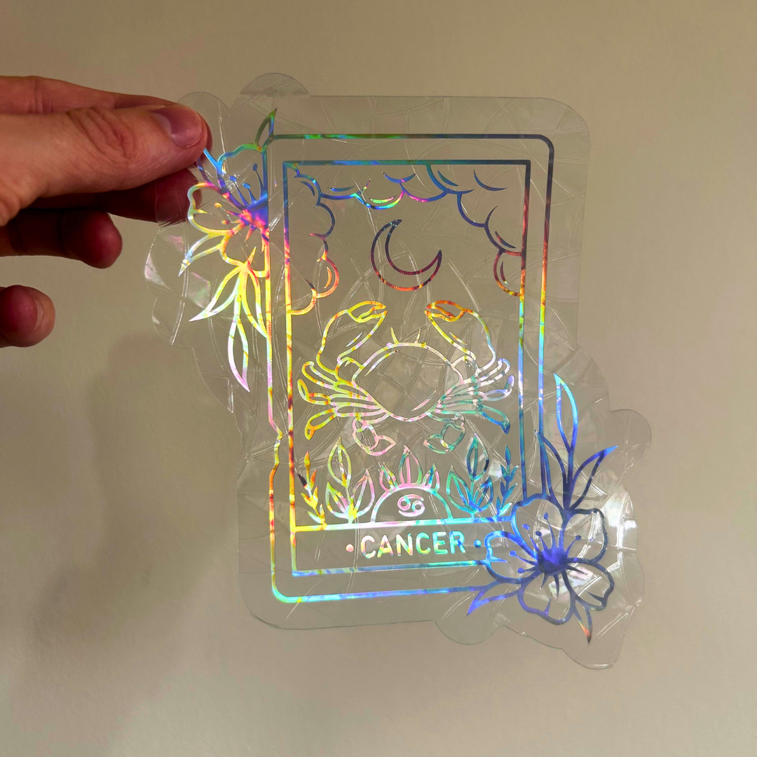 A hand holds a clear holographic sticker or decal featuring a Cancer zodiac card design with floral outlines and a moon symbol, shown against a plain beige wall.