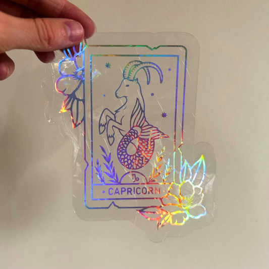 A hand holds a clear acrylic sticker featuring a Capricorn zodiac illustration with holographic rainbow foil accents and the text 'CAPRICORN' on a plain light background.