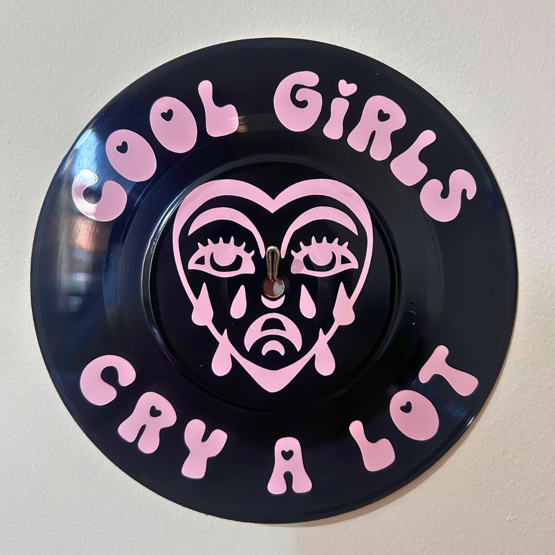 A circular black wall sign featuring pink text that reads 'COOL GIRLS CRY A LOT' and a pink heart-shaped face with tears in the center, mounted against a plain light wall.