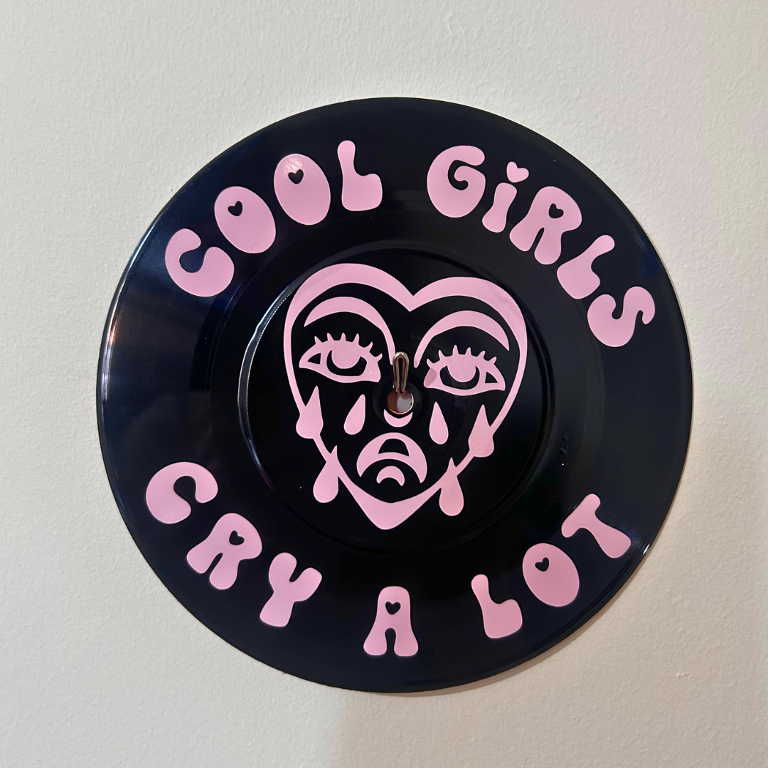 A circular black vinyl record  featuring the text 'COOL GIRLS CRY A LOT' in pink bubble lettering and a pink stylized face in the center, mounted on a plain off-white wall.