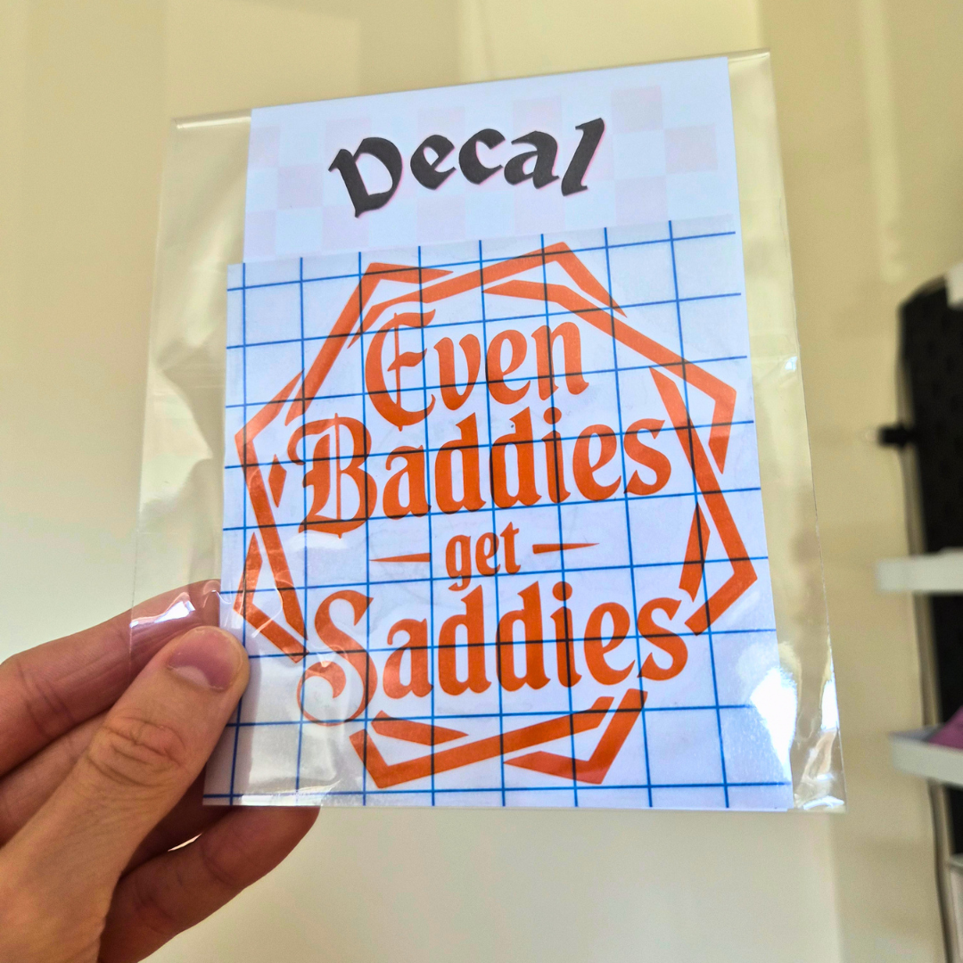 A hand holding a clear plastic-wrapped sticker or decal sheet with the text 'Even Baddies get Saddies' in orange lettering over a blue grid background.