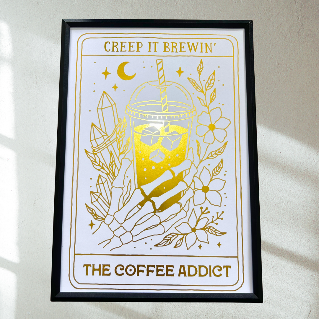 A framed art print featuring a gold coffee-themed illustration on a white background, hung on a light-colored wall with soft window shadows.