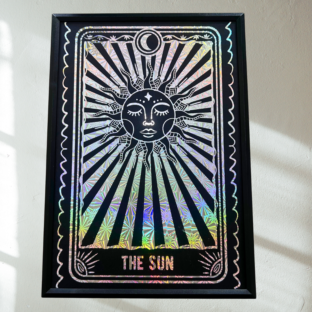 A framed tarot card art print featuring 'The Sun' with a black sun face and radiating rays over an iridescent holographic background, mounted on a light wall.