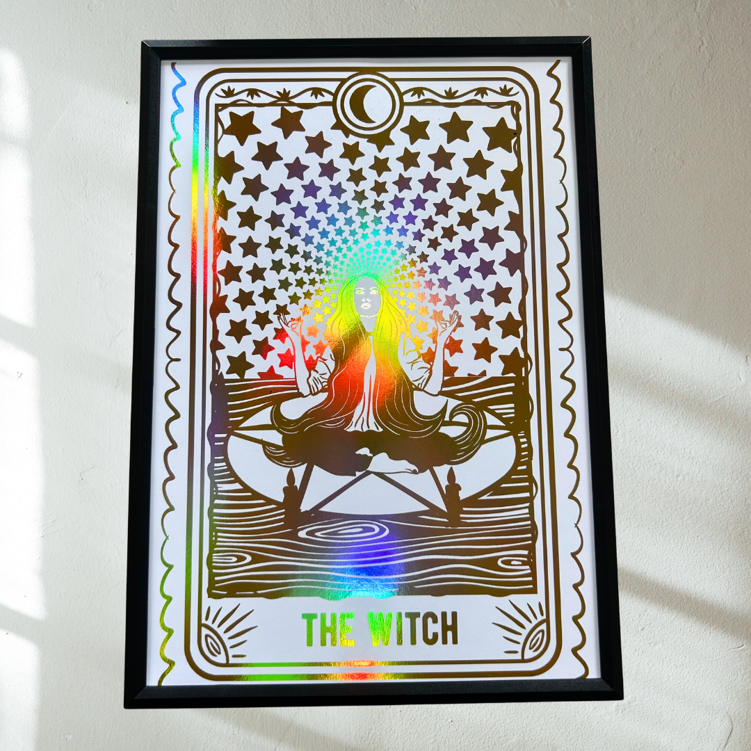 A framed tarot card art print titled 'THE WITCH' hanging on a light-colored wall. The design features a gold-toned witch figure surrounded by stars with a rainbow holographic glow, enclosed in a black frame.