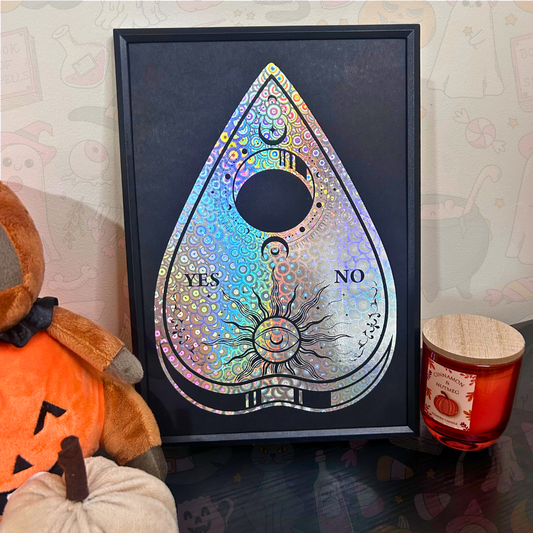 A framed black art print featuring a teardrop-shaped ouija board design with holographic silver foil details, set in a cozy home environment next to a plush toy and a candle.