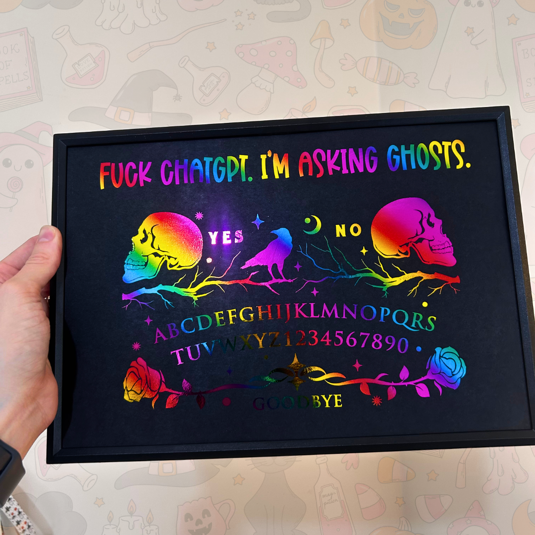 A hand holds a black-framed rainbow chalkboard art print featuring a skull, bird, roses, alphabet, and the phrase 'FUCK CHATGPT. I'M ASKING GHOSTS.