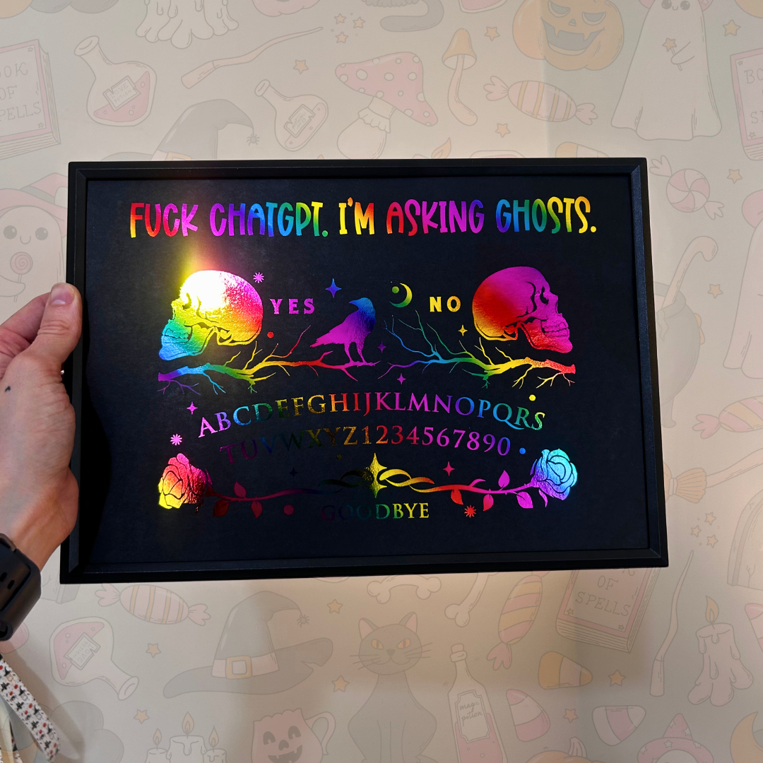A hand holds a black-framed glowing lightbox against a patterned wallpaper background. The lightbox displays a colorful, rainbow-lit Ouija board design with skulls, a raven, the words 'YES' and 'NO', alphabet letters, numbers, and the phrase 'FUCK CHATGPT'