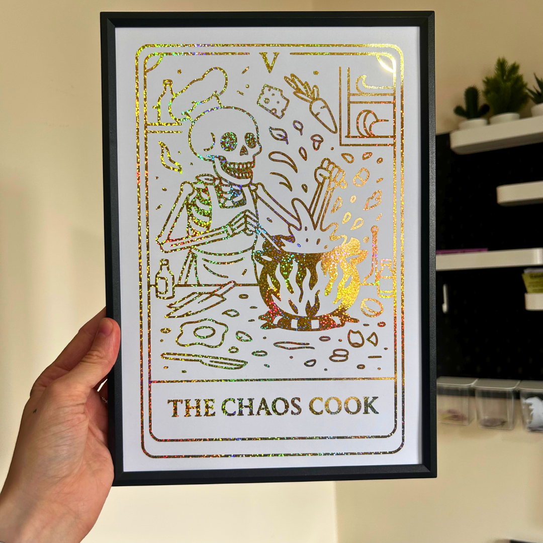 A hand holds a framed art print featuring a tarot-style skeleton cooking illustration with the text 'THE CHAOS COOK' at the bottom. The print has an iridescent gold foil texture and is housed in a black frame, set against an indoor background.