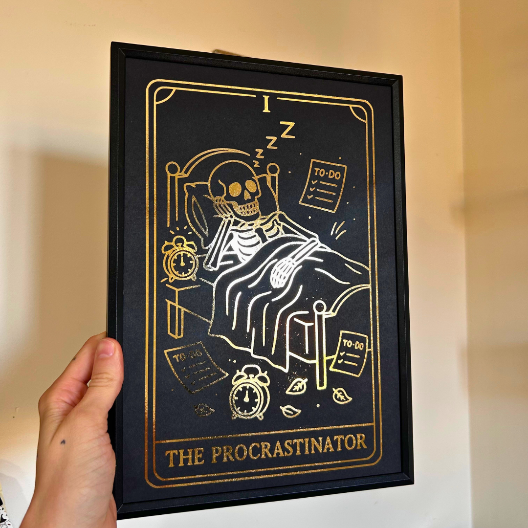 A hand holds a black-framed art print featuring a stylized tarot card illustration of a skeleton in bed with the text 'THE PROCRASTINATOR' in gold on a black background, against a beige wall.