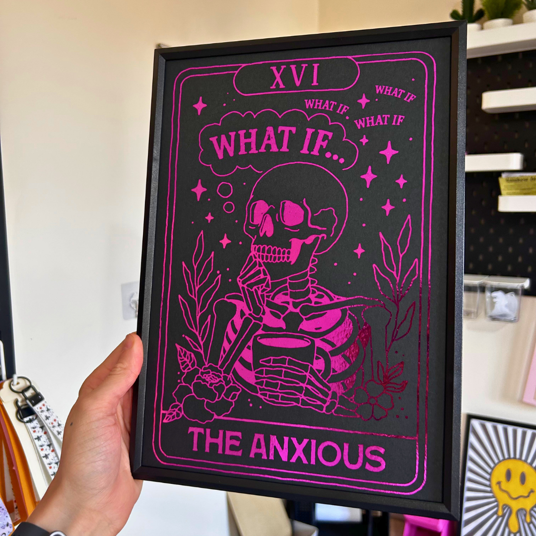 A hand holds a black-framed art print featuring a pink and black illustration of a skeleton wearing sunglasses, with the text 'XVI WHAT IF.