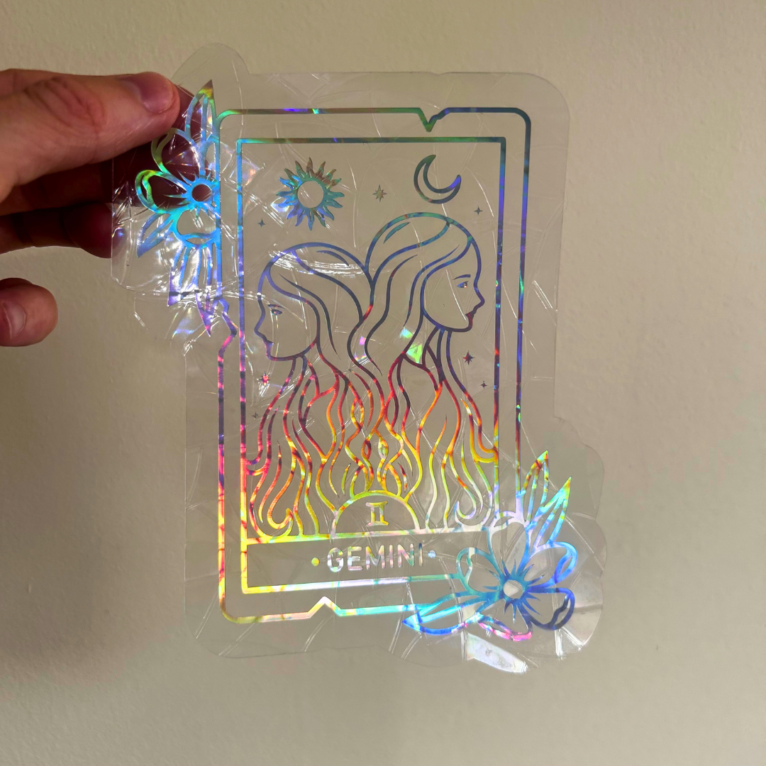 A hand holds a holographic tarot card sticker featuring the Gemini zodiac design, showing iridescent rainbow reflections against a plain beige wall.