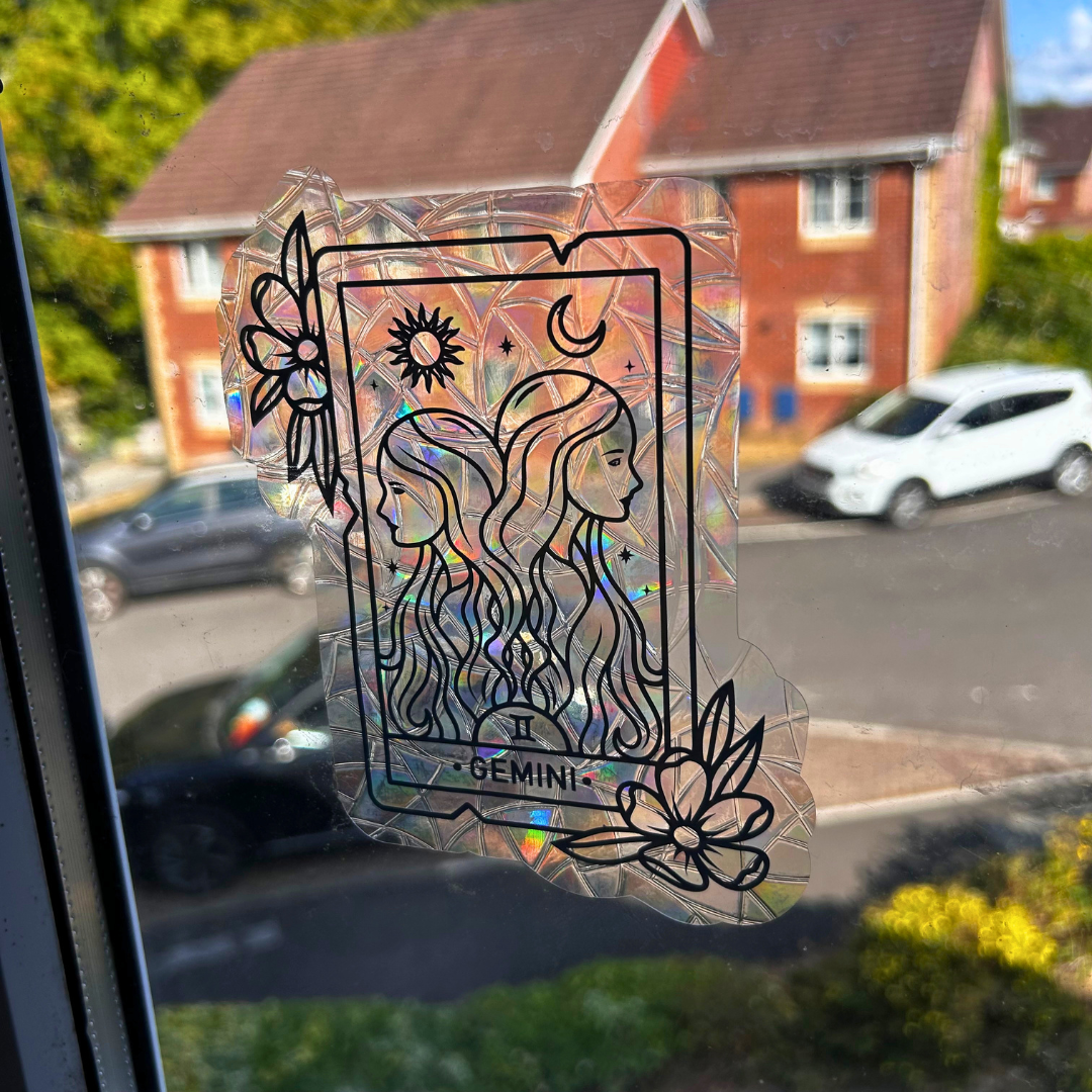 A holographic, crinkled clear sticker with a black tarot card design featuring two women and the word 'GEMINI', shown attached to a window with a blurred outdoor street scene behind it.