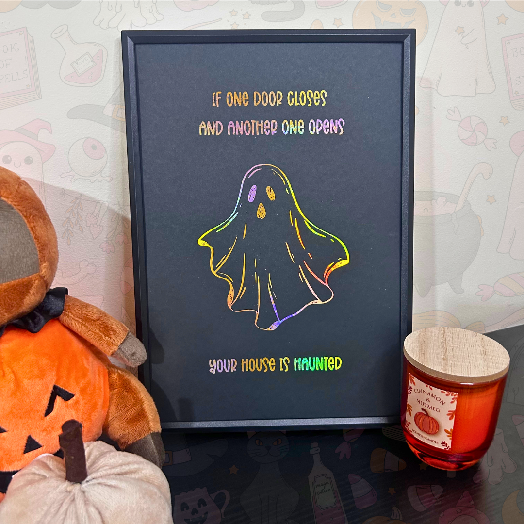 A framed Halloween art print with a dark charcoal background, featuring a colorful outline of a ghost and the text 'IF ONE DOOR CLOSSES AND ANOTHER ONE OPENS YOUR HOUSE IS HAUNTED'. The frame is styled on a dark reflective surface next to a plush pumpkin and a candle.