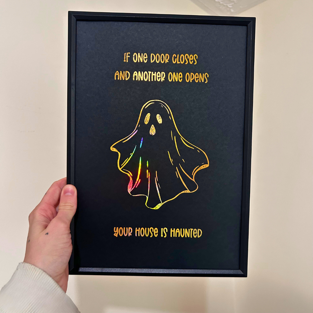 A hand holds a black framed art print featuring a holographic gold ghost and the text 'IF ONE DOOR CLOSES AND ANOTHER ONE OPENS YOUR HOUSE IS HAUNTED' against a beige wall.
