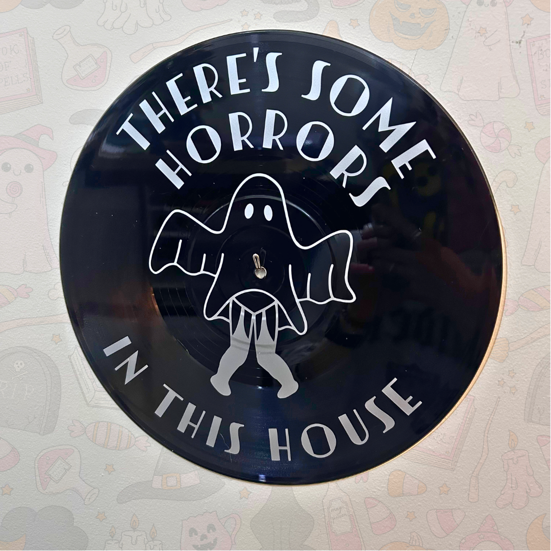 A circular black wall sign featuring a silver ghost illustration and the text 'THERE'S SOME HORRORS IN THIS HOUSE' on a patterned wall.