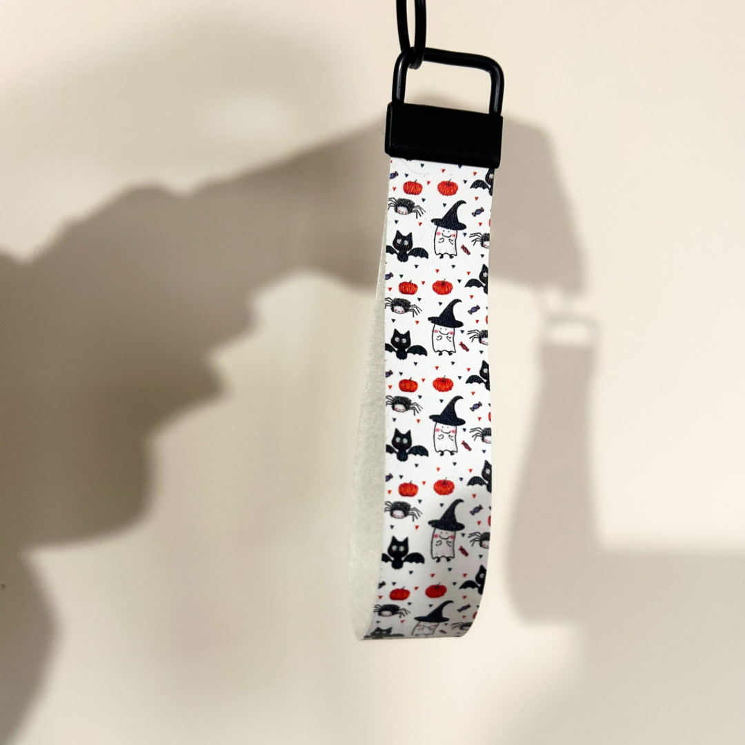 White wristlet with halloween design, cat with bat wings, pumpkin and ghosts, with black hardware 