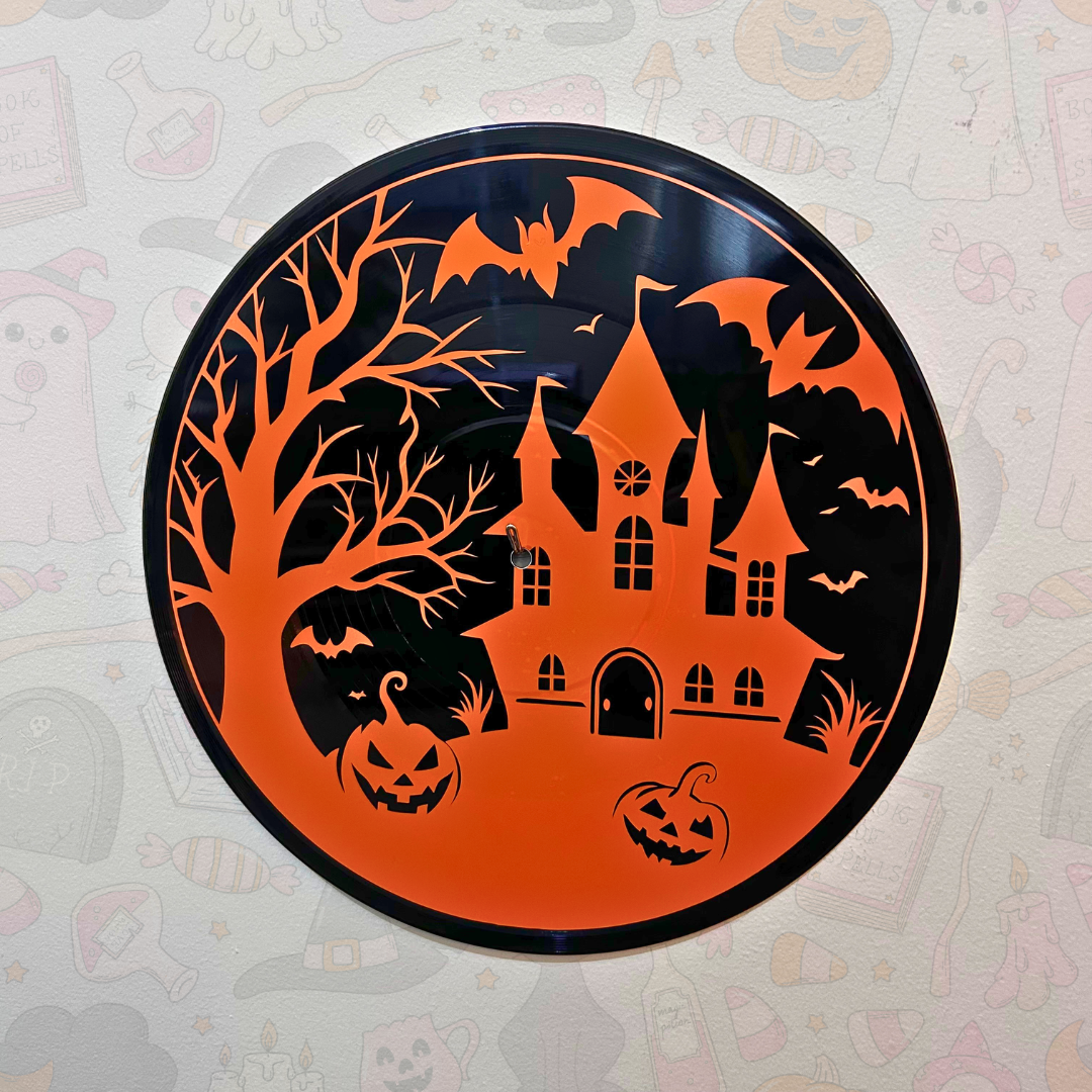 A circular Halloween wall art piece featuring a black silhouette of a haunted house, bare tree, bats, pumpkins, and spooky trees on an orange background, mounted against a light patterned wall.