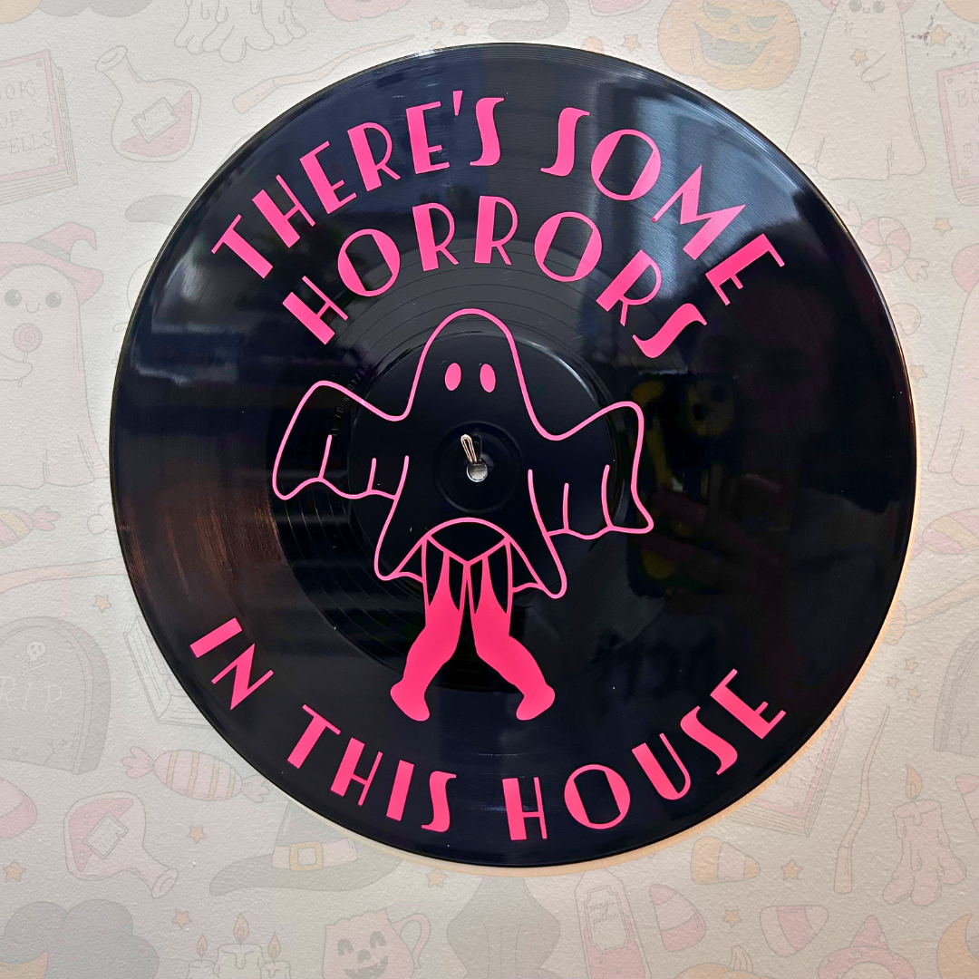 A circular black vinyl record wall art piece featuring pink text that reads 'THERE'S SOME HORRORS IN THIS HOUSE' and a pink ghost illustration in the center, mounted against a light patterned wall.