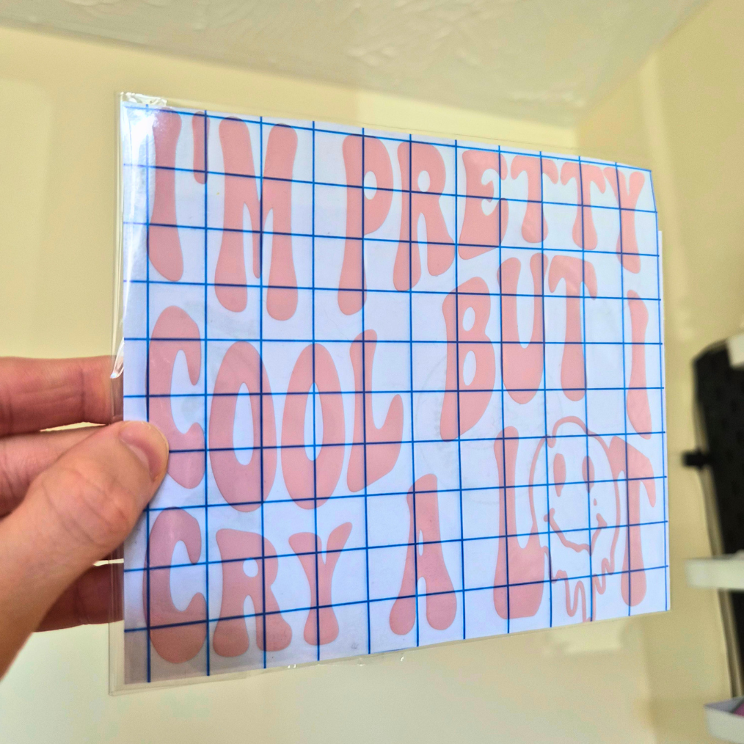 A hand holds a square sticker sheet featuring pink bubble letters on a white grid background with the text 'I'M PRETTY COOL BUT I CRY A LOT' and a smiley face graphic.