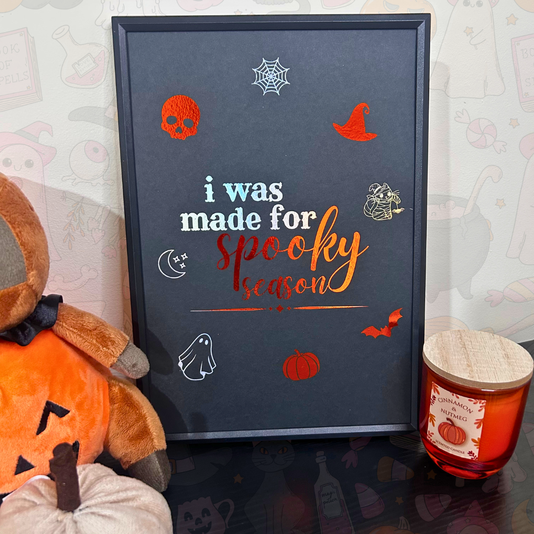 A framed Halloween-themed art print with the text 'i was made for spooky season' in white and orange lettering, surrounded by illustrations of a skull, witch hat, spiderweb, ghost, pumpkin, bat, and moon. The framed print is styled on a tabletop next to a plush pumpkin, a teddy bear, and a candle.