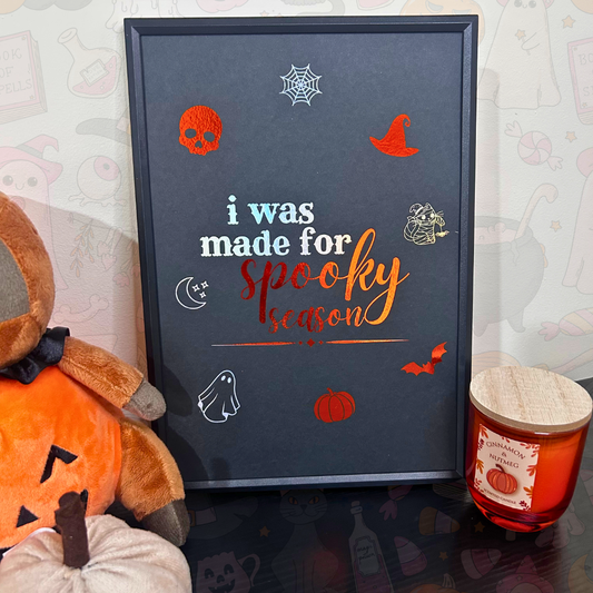 A framed Halloween-themed art print with the text 'i was made for spooky season' in white and orange lettering, surrounded by illustrations of a skull, witch hat, spiderweb, ghost, pumpkin, bat, and moon. The framed print is styled on a tabletop next to a plush pumpkin, a teddy bear, and a candle.
