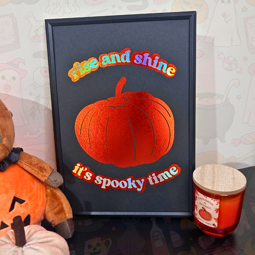 A framed Halloween art print featuring a red-orange pumpkin and the text 'Rise and shine it's spooky time' in colorful letters, styled on a tabletop next to a teddy bear and a candle.
