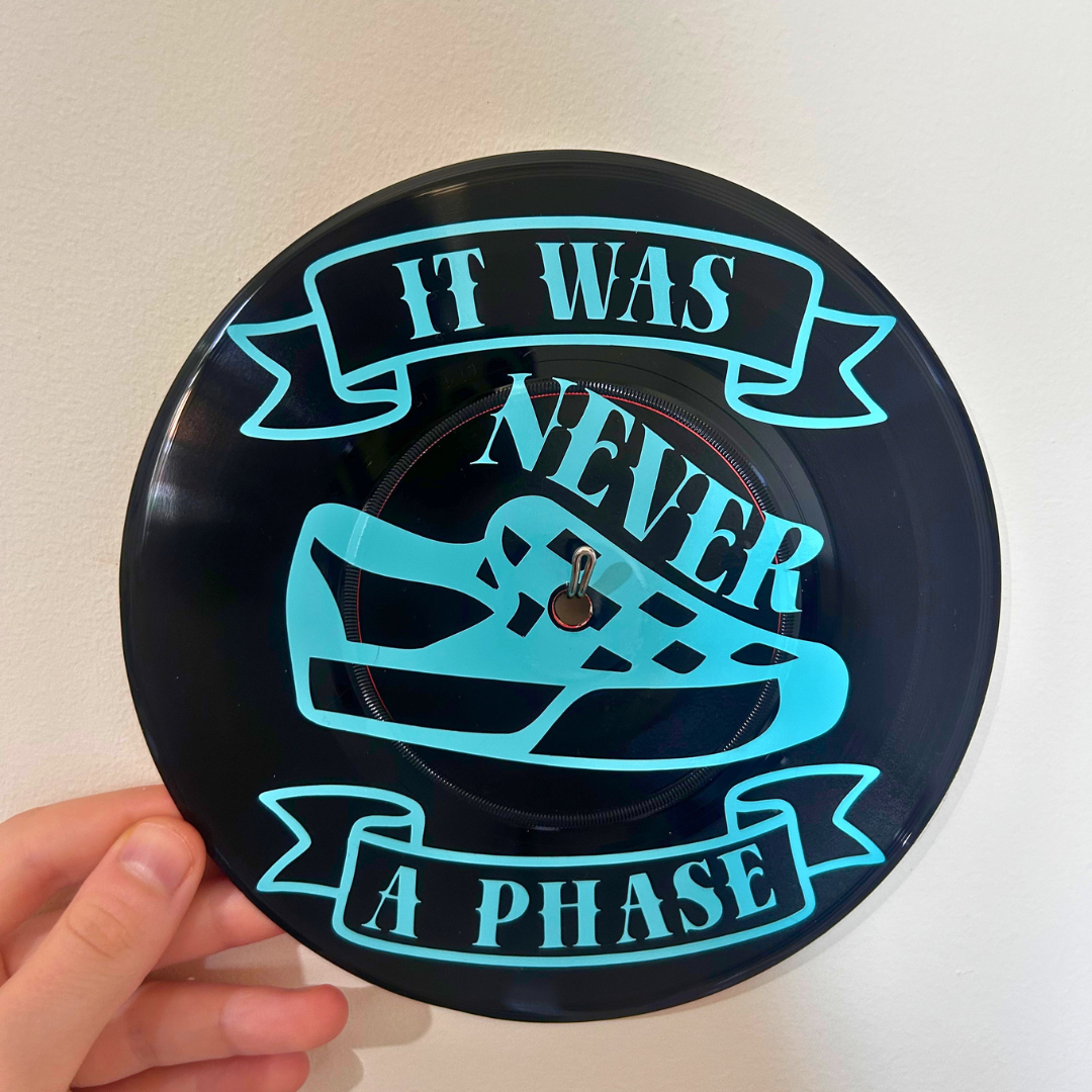 ,A circular black vinyl record wall decor  held by a hand, featuring a graphic of a sneaker and banners with the text 'IT WAS NEVER A PHASE'.