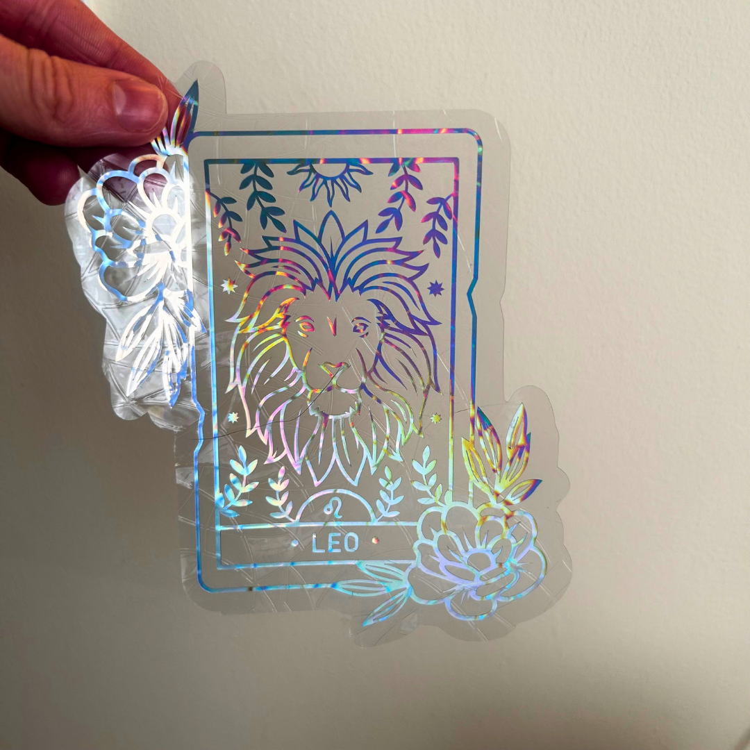 A hand is holding a clear vinyl sticker against a plain wall. The sticker features a colorful holographic design of a lion card with floral details and the word 'LEO' at the bottom.