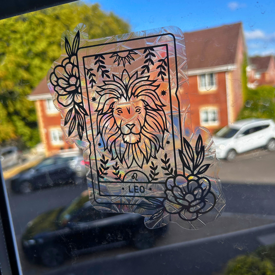 A clear, holographic vinyl decal of the Leo tarot card applied to a window, with a blurred street scene of houses and cars in the background.