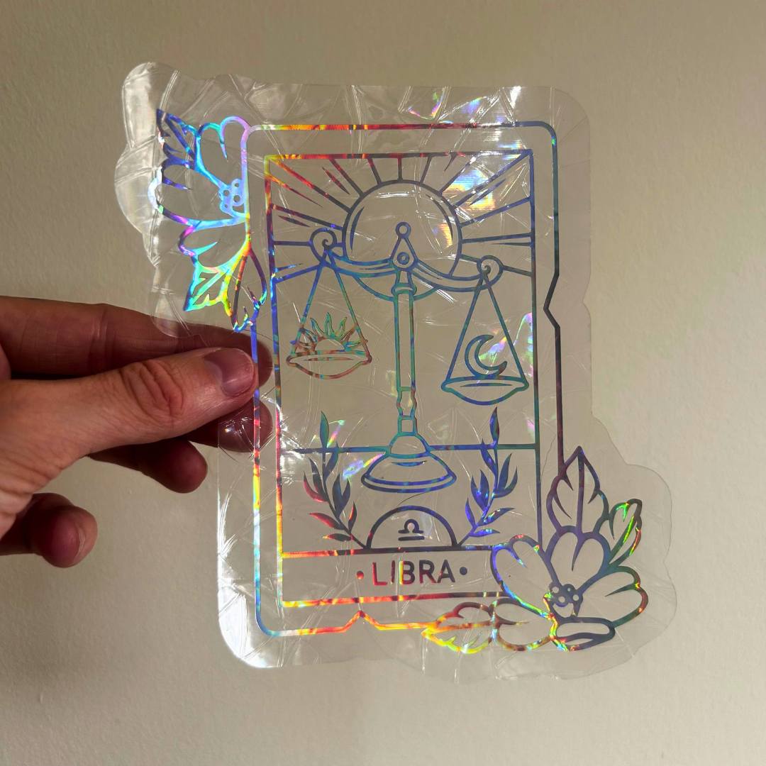 A hand holds a clear acrylic sticker featuring a Libra tarot card design with holographic rainbow foil accents, shown against a plain beige wall.