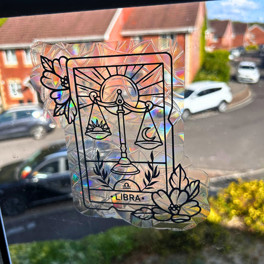 A holographic, iridescent sticker of the Libra tarot card design, featuring a black line art illustration of the scales of justice, sun, moon, and flowers, applied to a window with a blurred suburban street background.